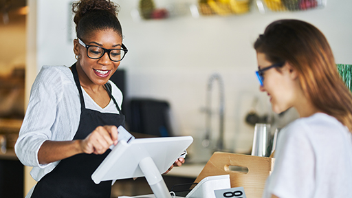 A female employee stands behind a cafe counter facing a female guest. The employee swipes the customer's credit card. 