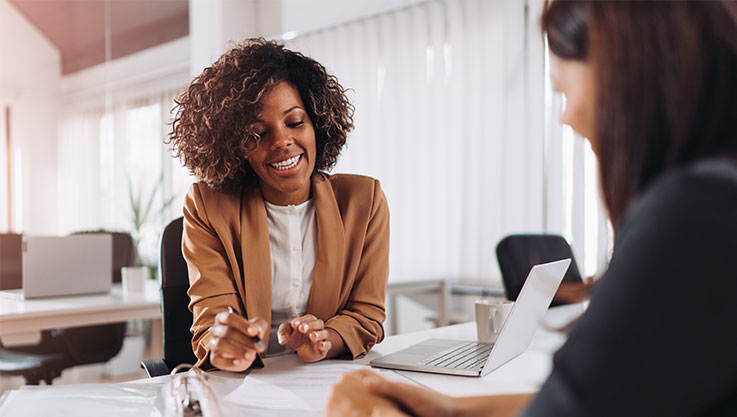 Two women sit across a table from each other. On the table are papers and binders. 