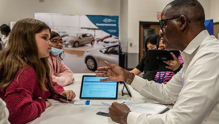 An adult volunteer speaks with a student at a table. In between them is a tablet with a budgeting exercise.