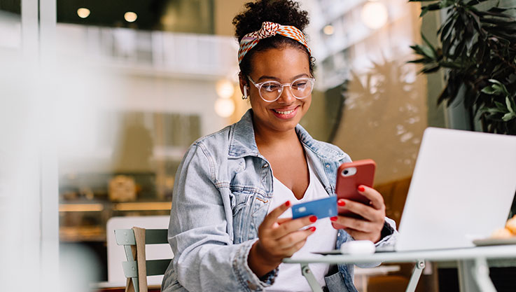 A person sits at a cafe table holding a credit card and a phone. 