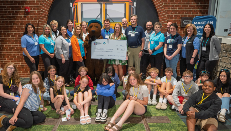 Adults, students, and a mascot stand for a photo with a large promotional check. 