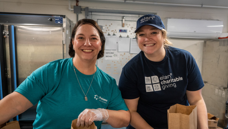 Two volunteers stand together, smiling, in a commerical kitchen packaging food. 