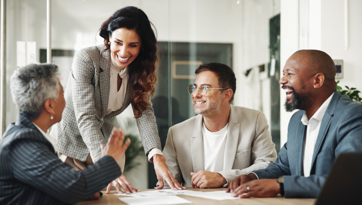 Four business people gather in a conference room. There is paperwork on the counter and they are smiling in discussion. 