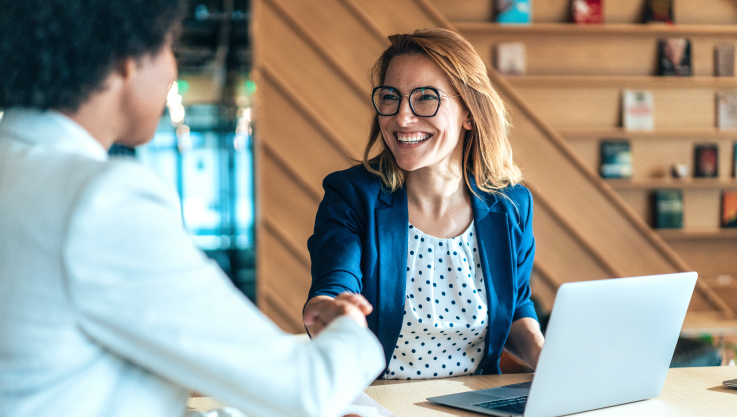 Two women sit at a conference table shaking hands. 