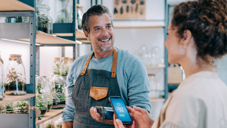 A store owner holds a payment terminal in his hand. A customer holds her phone over the terminal to pay. 