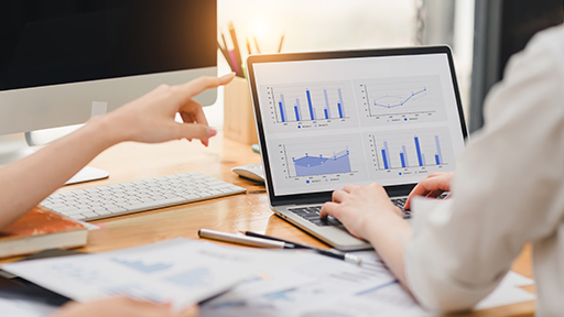 A person sits at a desk with their laptop open. There are papers covering the table and the screen shows graphs. 
