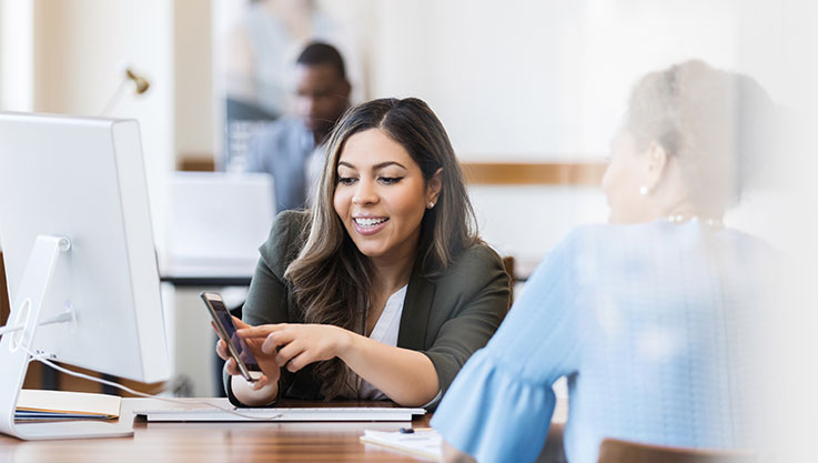 A customer sits at a desk with a staff member. They are working together on a mobile device. 